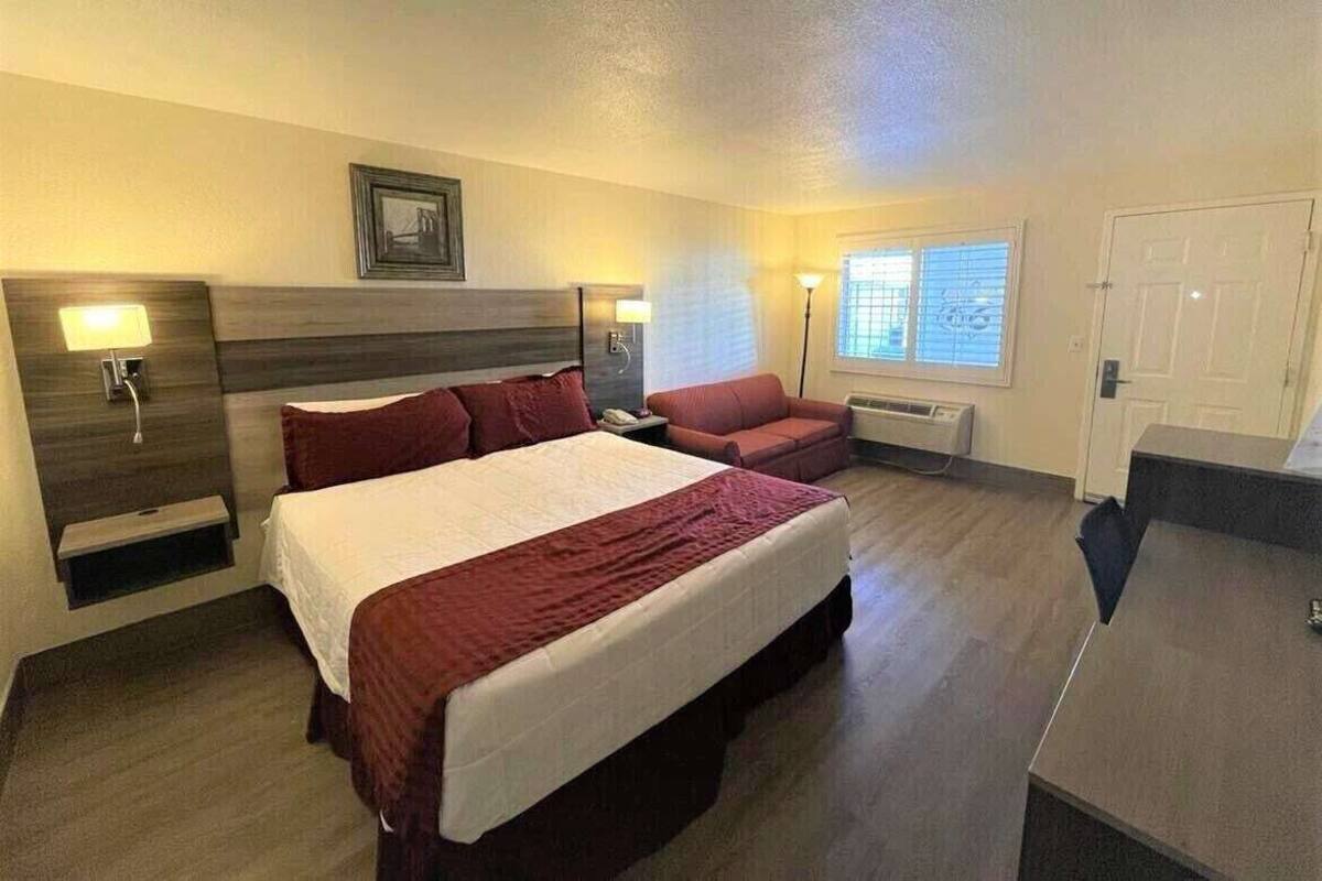 Ein Hotelzimmer mit einem großen Bett, einem Schreibtisch, einem Sessel, einer Couch, einem Nachttisch mit Lampe, einem Bild an der Wand und einem Fenster mit Jalousien.