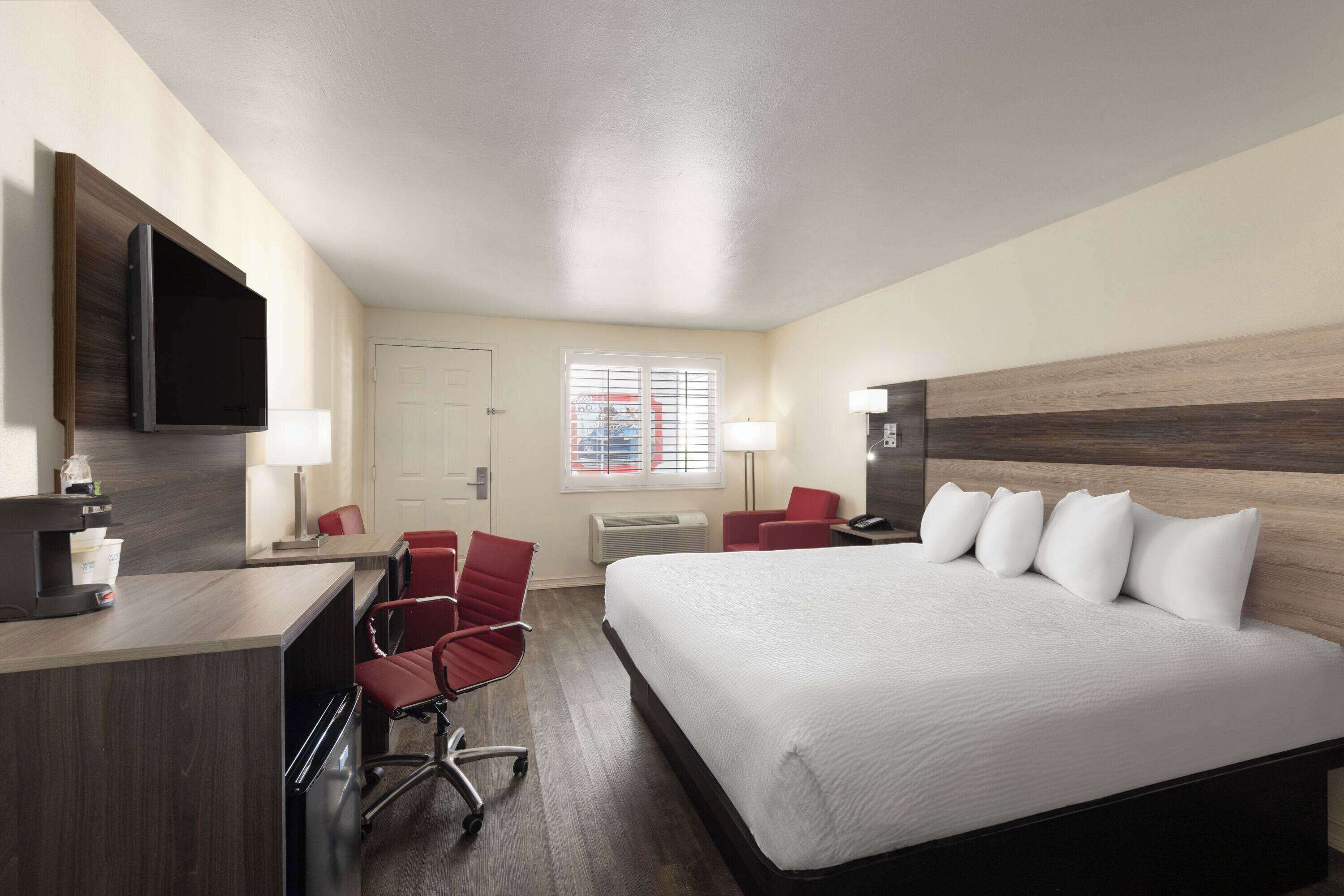 Quarto de hotel com cama, escrivaninha, cadeiras, televisão e janela com persianas.