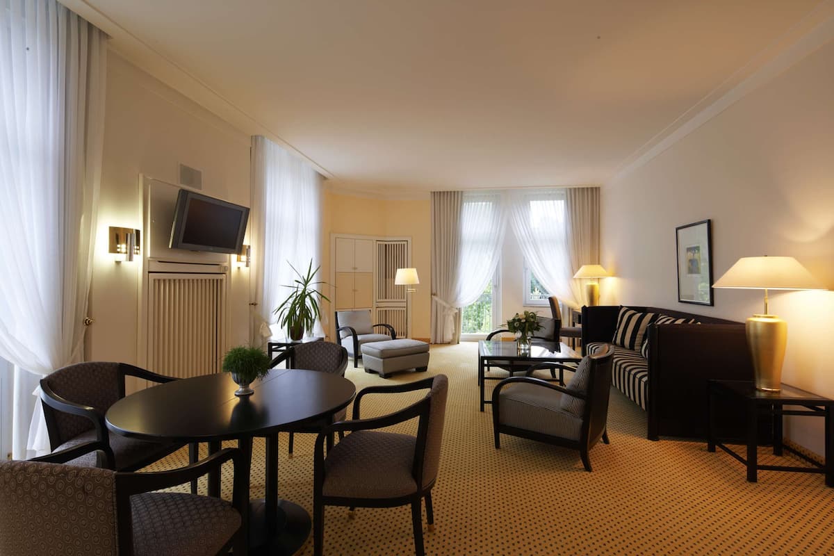 Une chambre d’hôtel comprenant un coin repas, un canapé, une télévision et une grande fenêtre avec des rideaux.