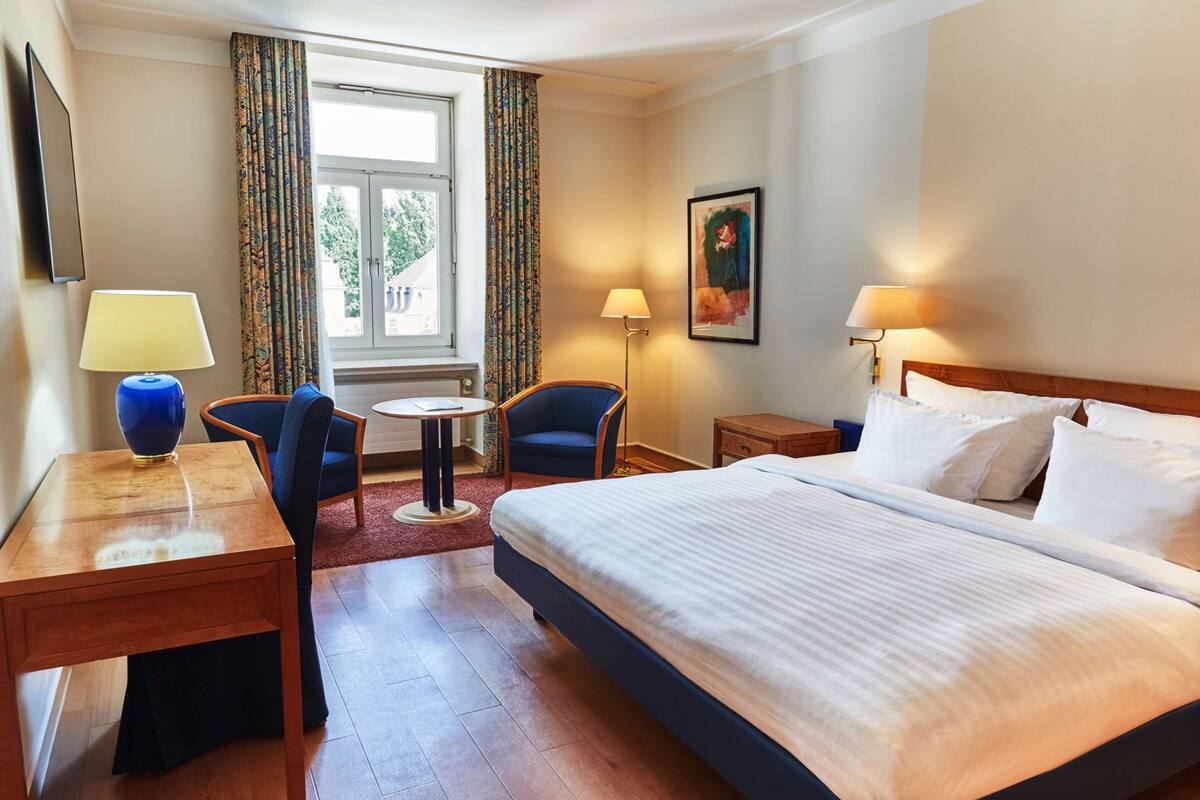 Une chambre d’hôtel avec un grand lit, un bureau avec une lampe, une chaise, une petite table et une fenêtre avec des rideaux.