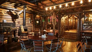 Een rustiek blokhutinterieur met een houten eettafel, metalen stoelen, een open haard en diverse decoraties.