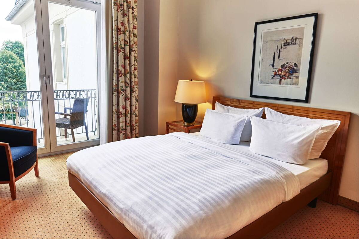 Une chambre d’hôtel avec un lit, une chaise, une table de chevet avec une lampe, un balcon avec une chaise et un tableau encadré au mur.