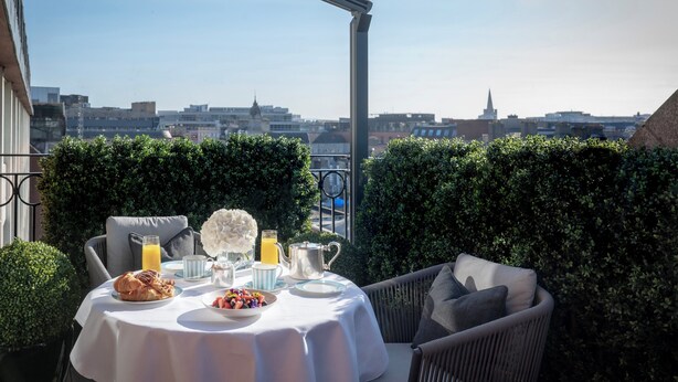 Mesa lista para el desayuno con croissants, fruta y jugo, con vista a un paisaje urbano.