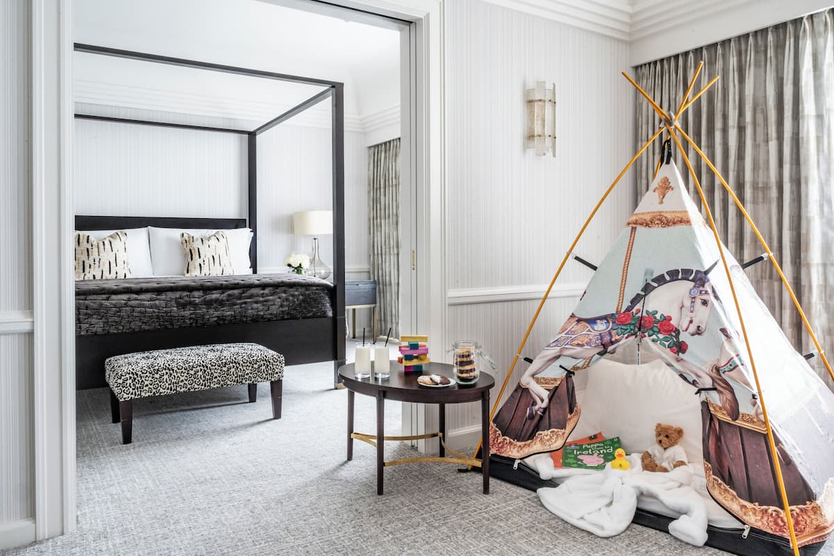 Un dormitorio con cama con dosel, un banco, una mesita con libros y un oso de peluche, y una tienda tipi.