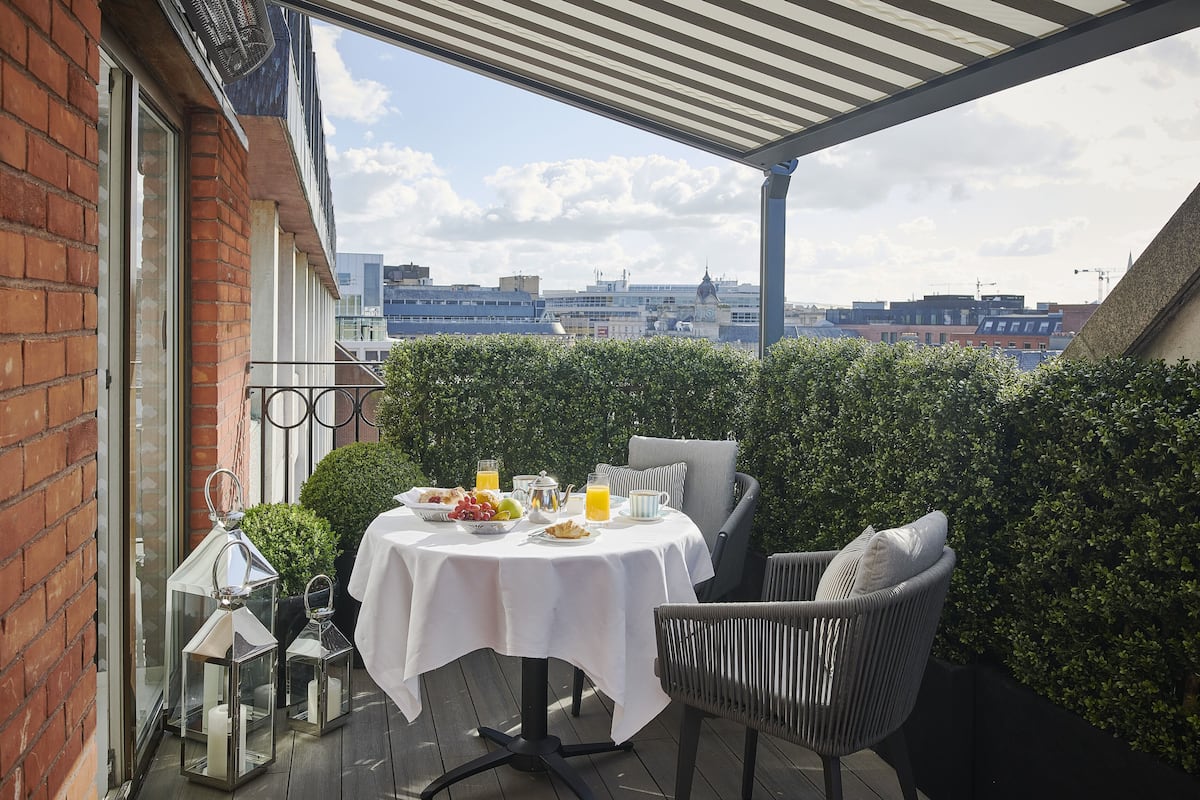 Terraza en la azotea con una mesa dispuesta para el desayuno, rodeada de vegetación y con vistas a un paisaje urbano.