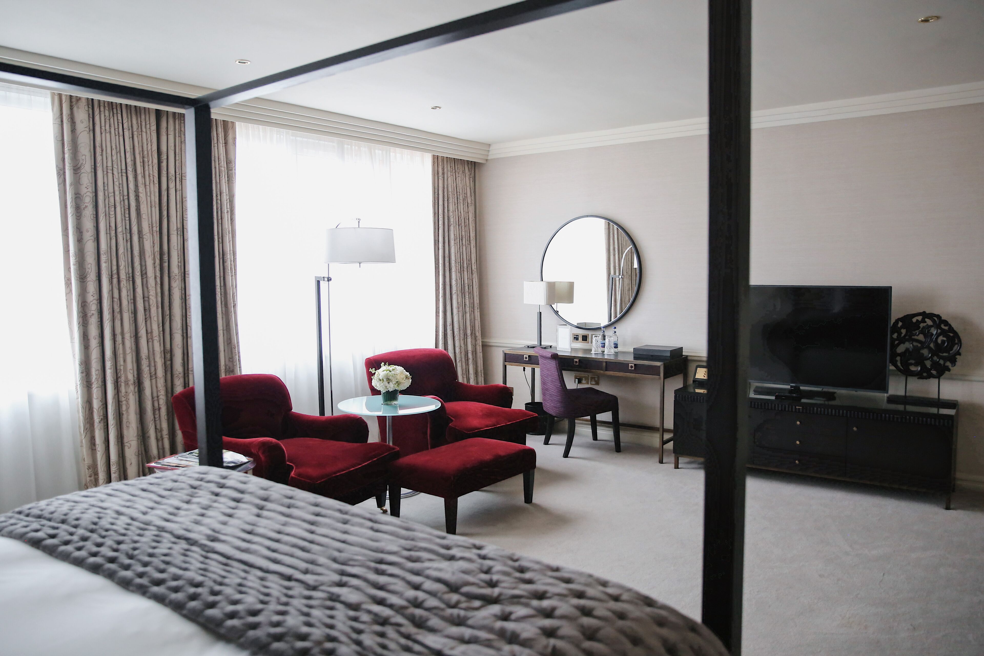 A hotel room with a large bed, a desk with a computer, a TV, and two red armchairs.
