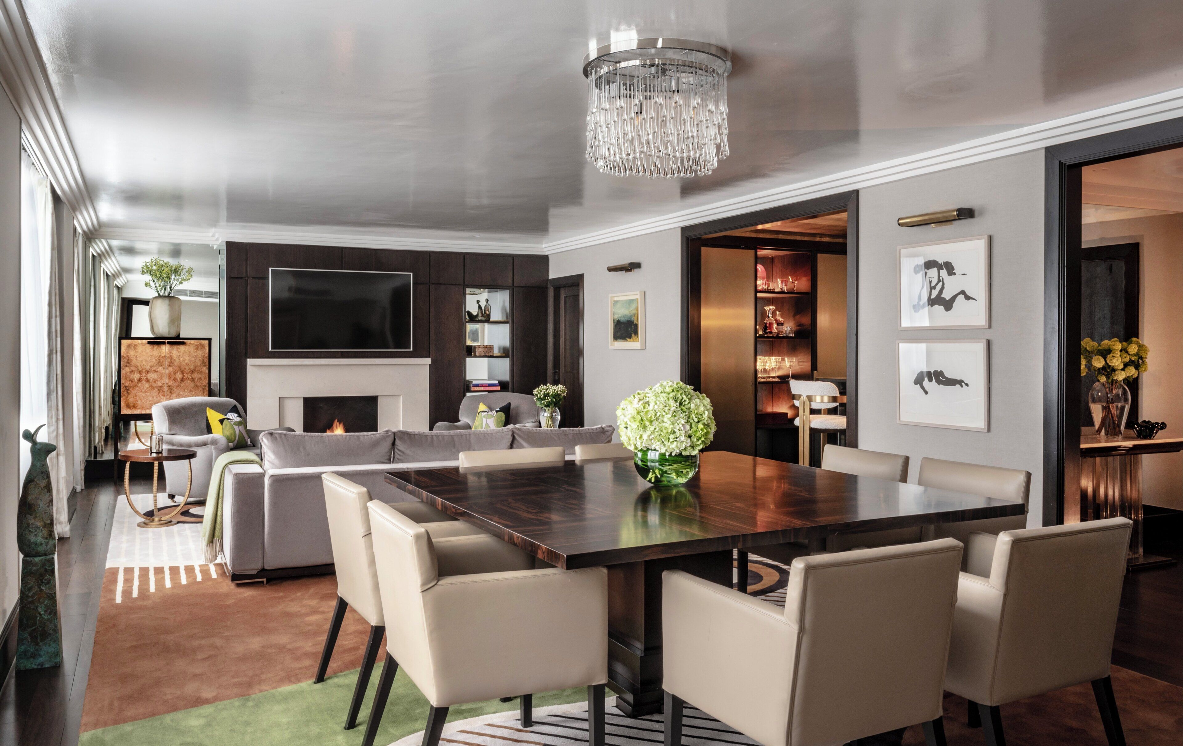 A modern living room with a large dark wood dining table, beige chairs, a fireplace, a flat-screen TV, and a chandelier.