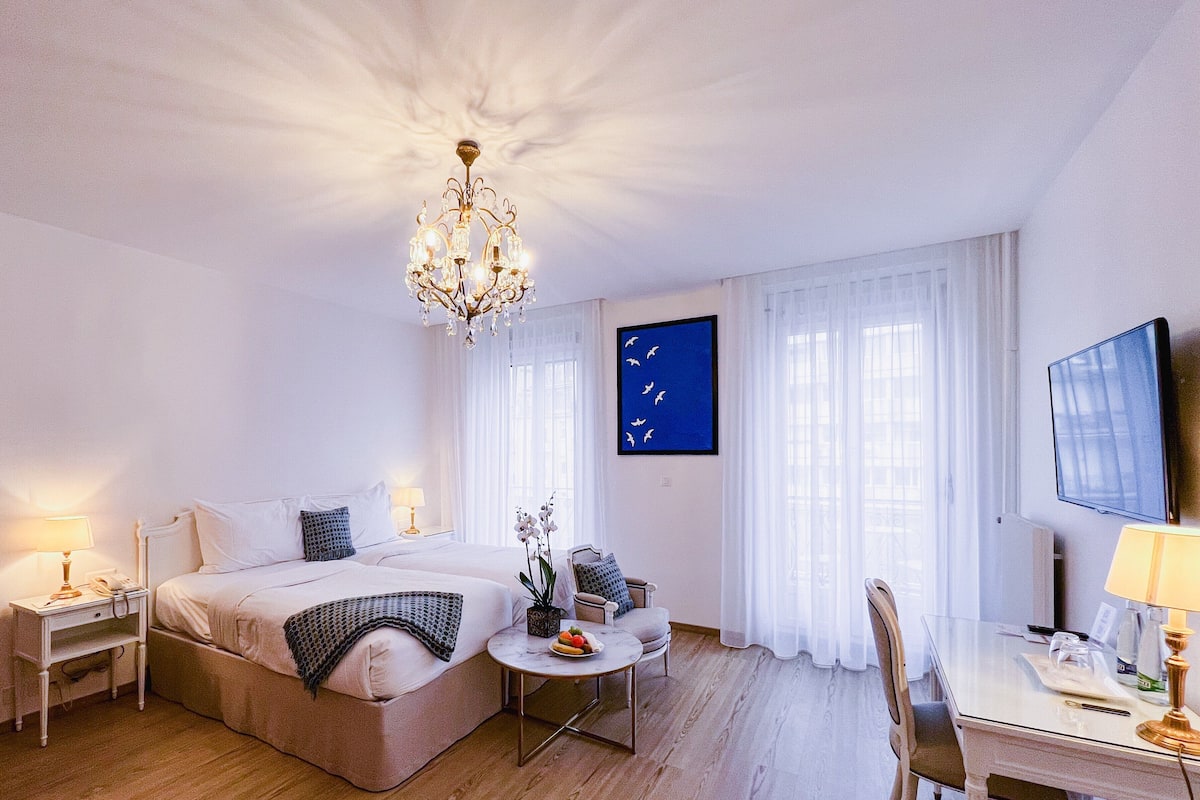 Habitación de hotel con una cama grande, un escritorio, una silla, una mesita, un televisor y una araña.
