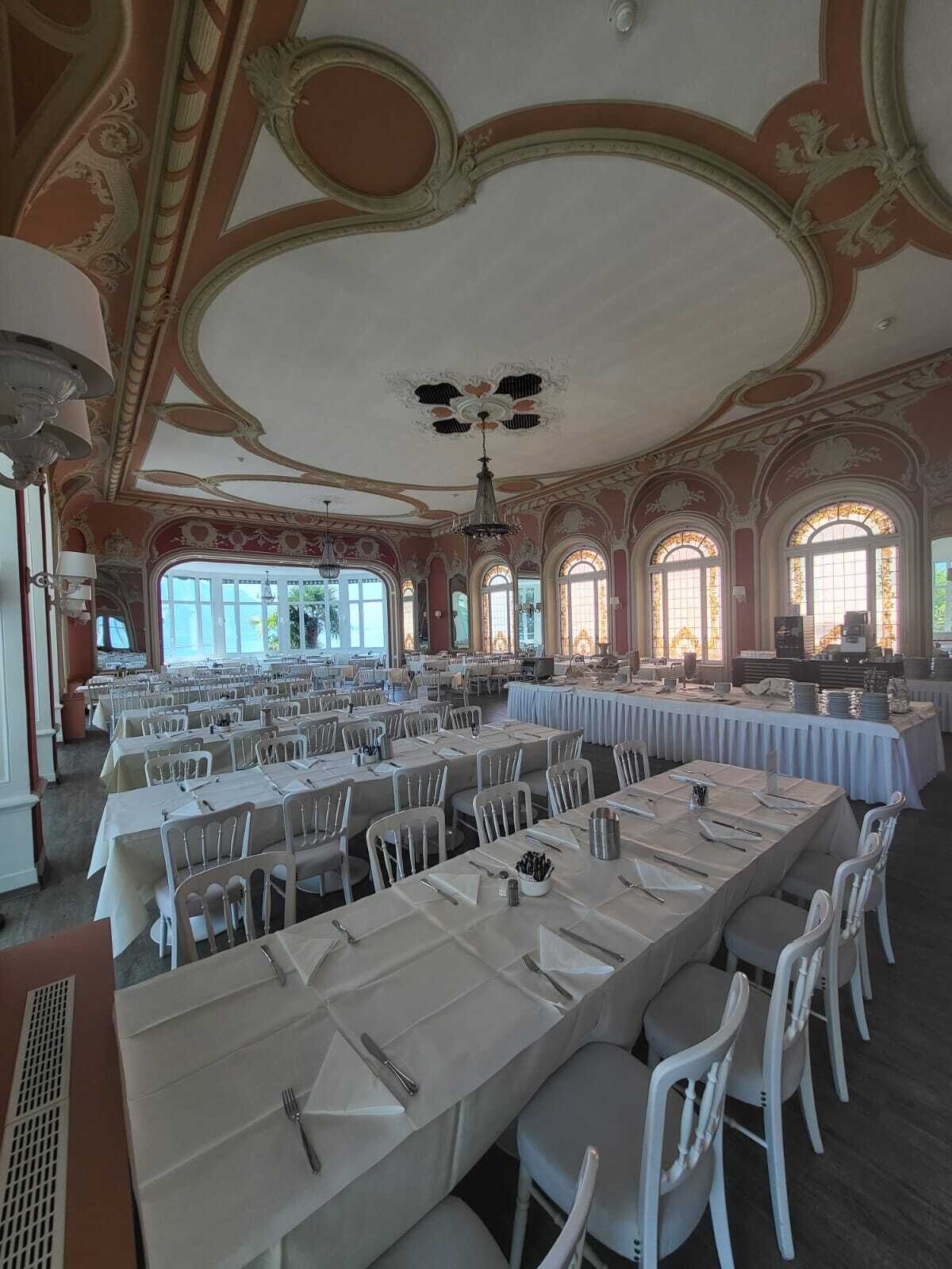 A banquet hall with long tables, white chairs, and ornate ceiling decorations.