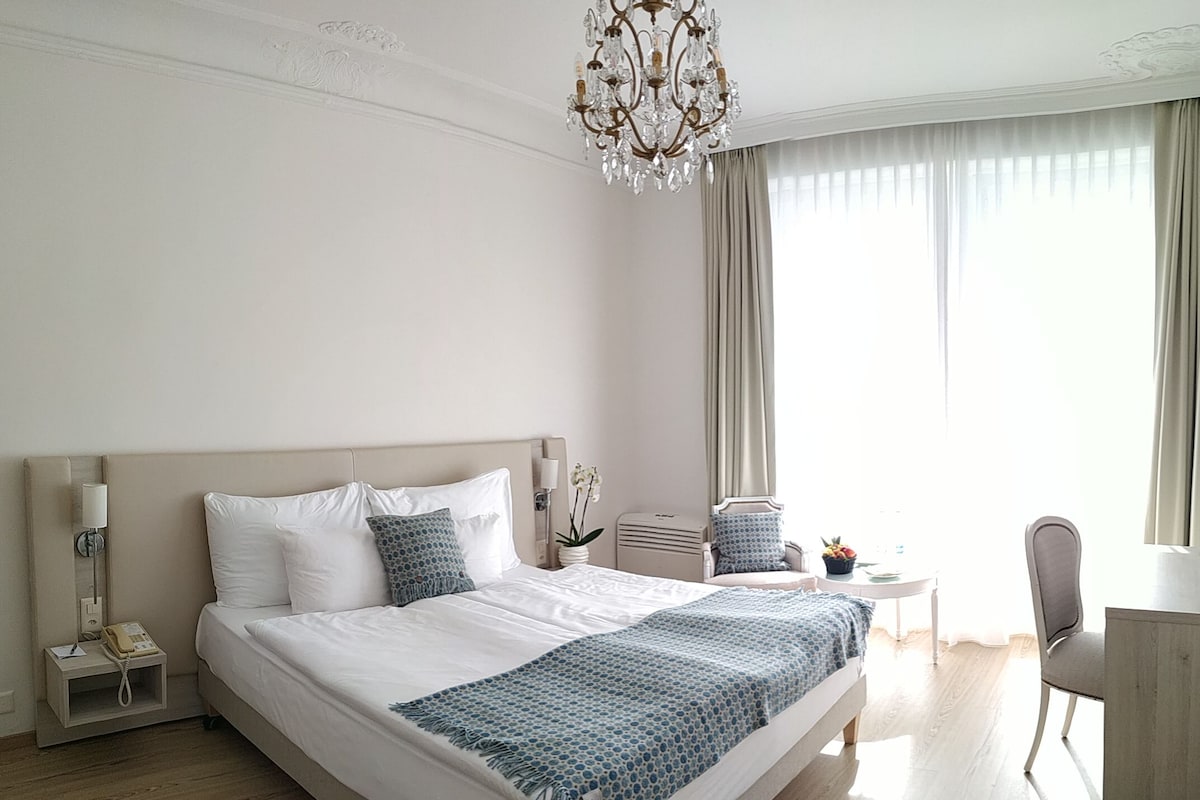 Een hotelkamer met een groot bed, nachtkastjes, een bureau, een stoel, een kroonluchter en een raam met gordijnen.