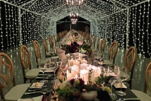 A long dining table with candles, glassware, and floral centerpieces, set in a room with a geometric ceiling design and wooden chairs.