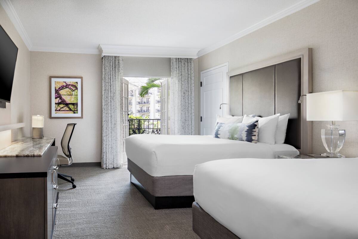 Une chambre d’hôtel avec deux lits, un bureau, une chaise, une télévision et une fenêtre donnant sur des immeubles et des espaces verts.