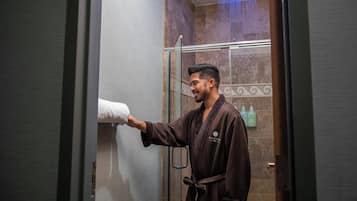 A person in a robe holding a towel in a bathroom with a shower and toiletries on the wall.