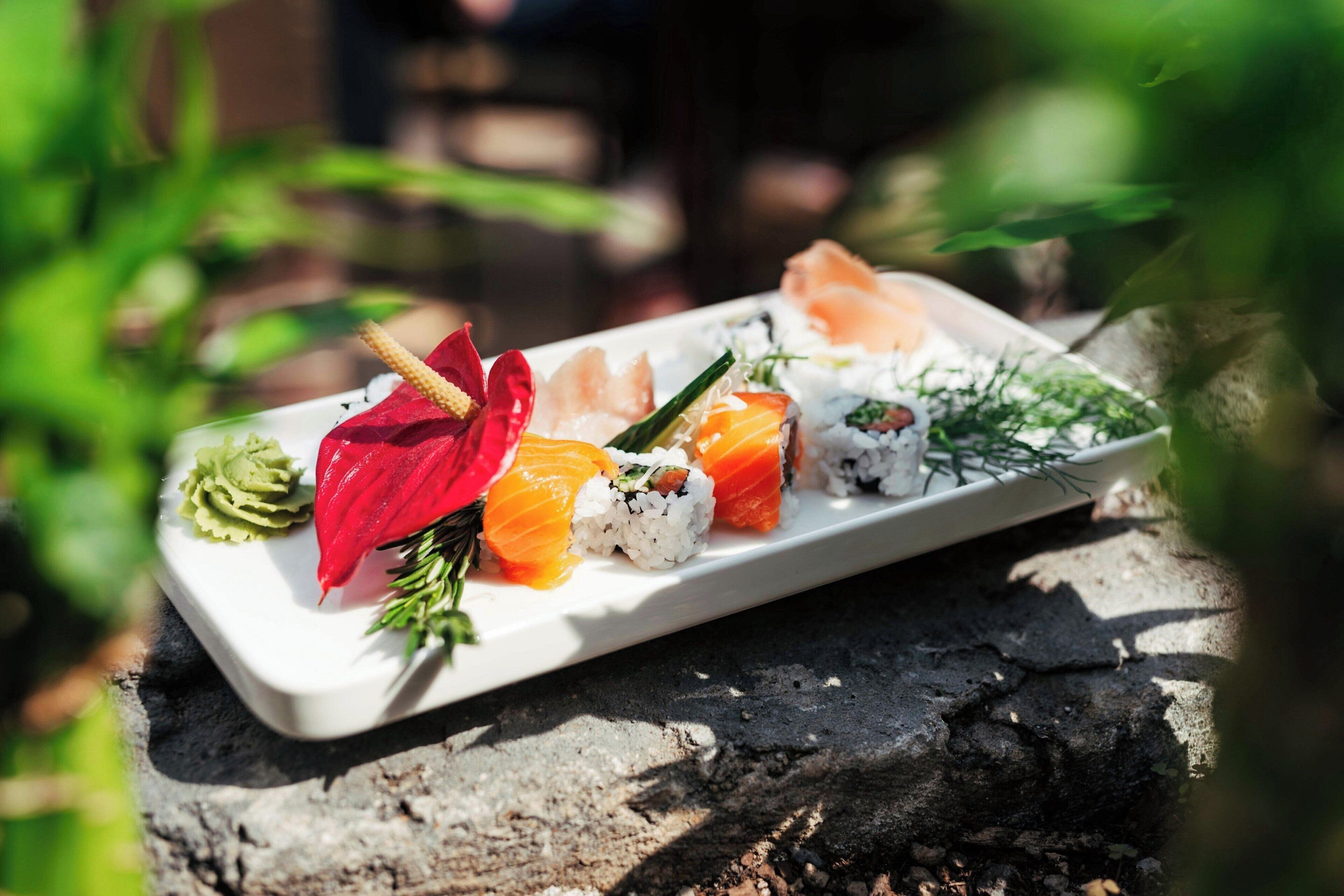 A sushi platter on a white plate with various types of sushi, including salmon and avocado, garnished with green leaves and a red flower.
