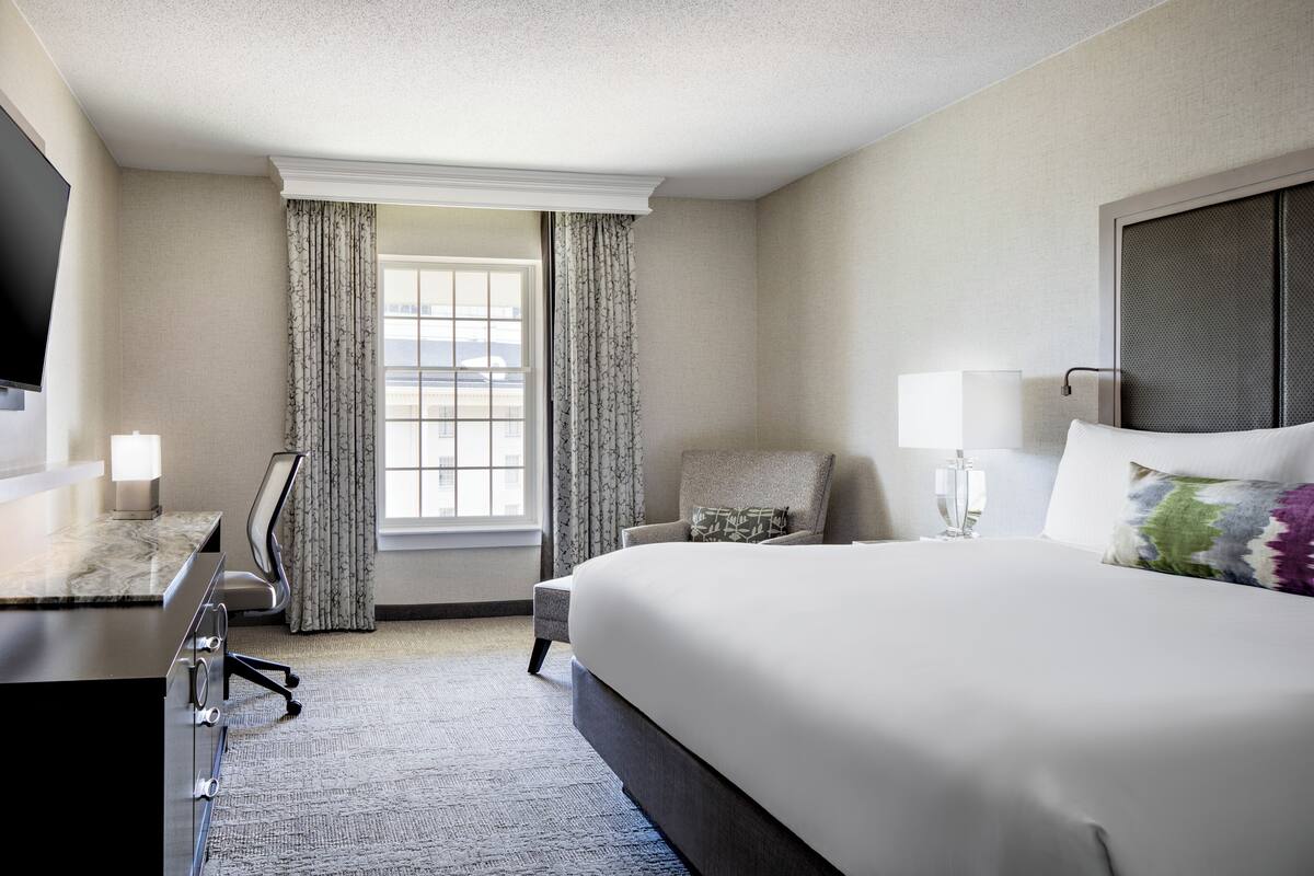 Une chambre d’hôtel avec un grand lit, un bureau avec une chaise, une télévision et une fenêtre avec des rideaux.