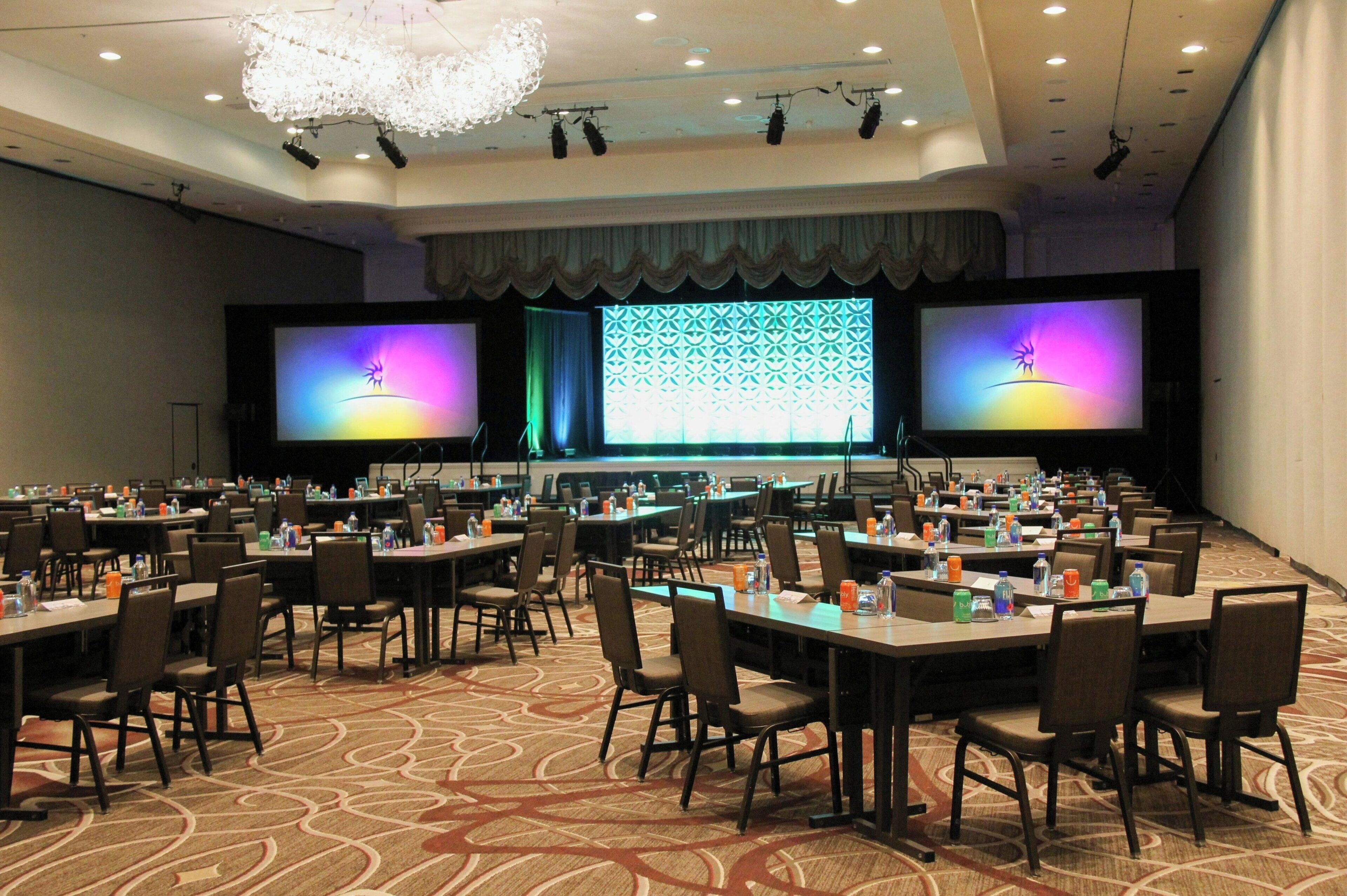A conference room with multiple tables and chairs, a large screen, and a stage with lighting.