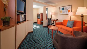 A hotel room with a sofa, armchair, TV, desk, lamp, and a decorative plant.