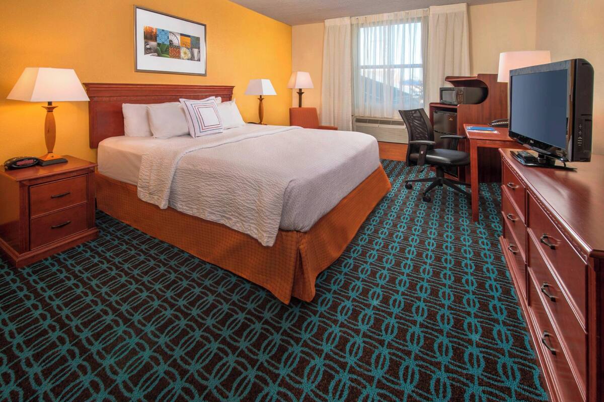Une chambre d’hôtel avec un grand lit, deux tables de chevet avec des lampes, un bureau avec un ordinateur, une télévision et une moquette à motifs.