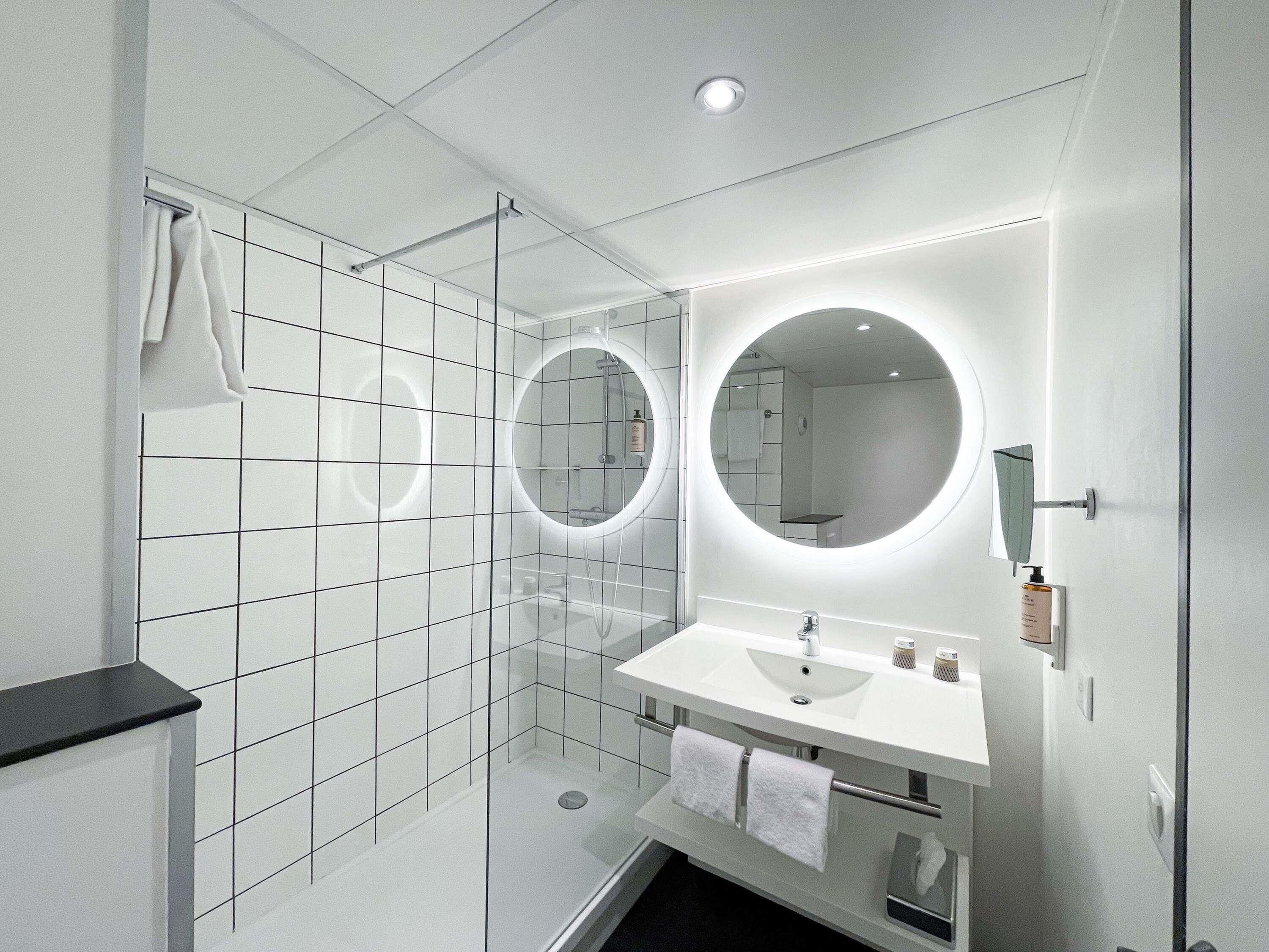 Een moderne badkamer met een witte betegelde douche, een ronde spiegel en een witte wastafel.