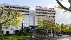 Un hotel con fachada moderna, un cartel llamativo que dice "Mercure Hotel" y un área exterior bien cuidada con árboles y cielo despejado.