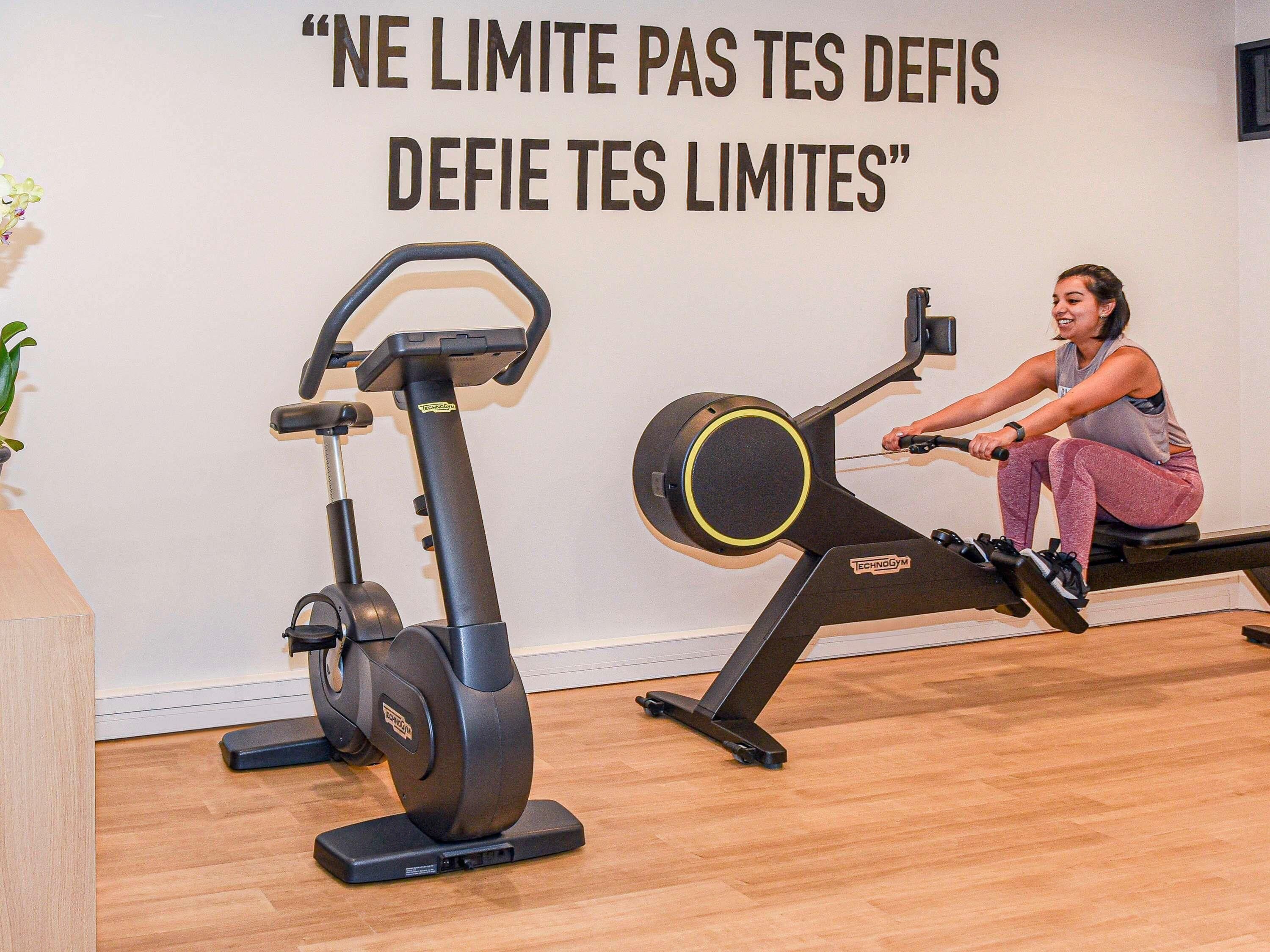 Une femme s’entraîne sur un appareil de rame dans une salle de sport, et un message motivant est affiché sur le mur.