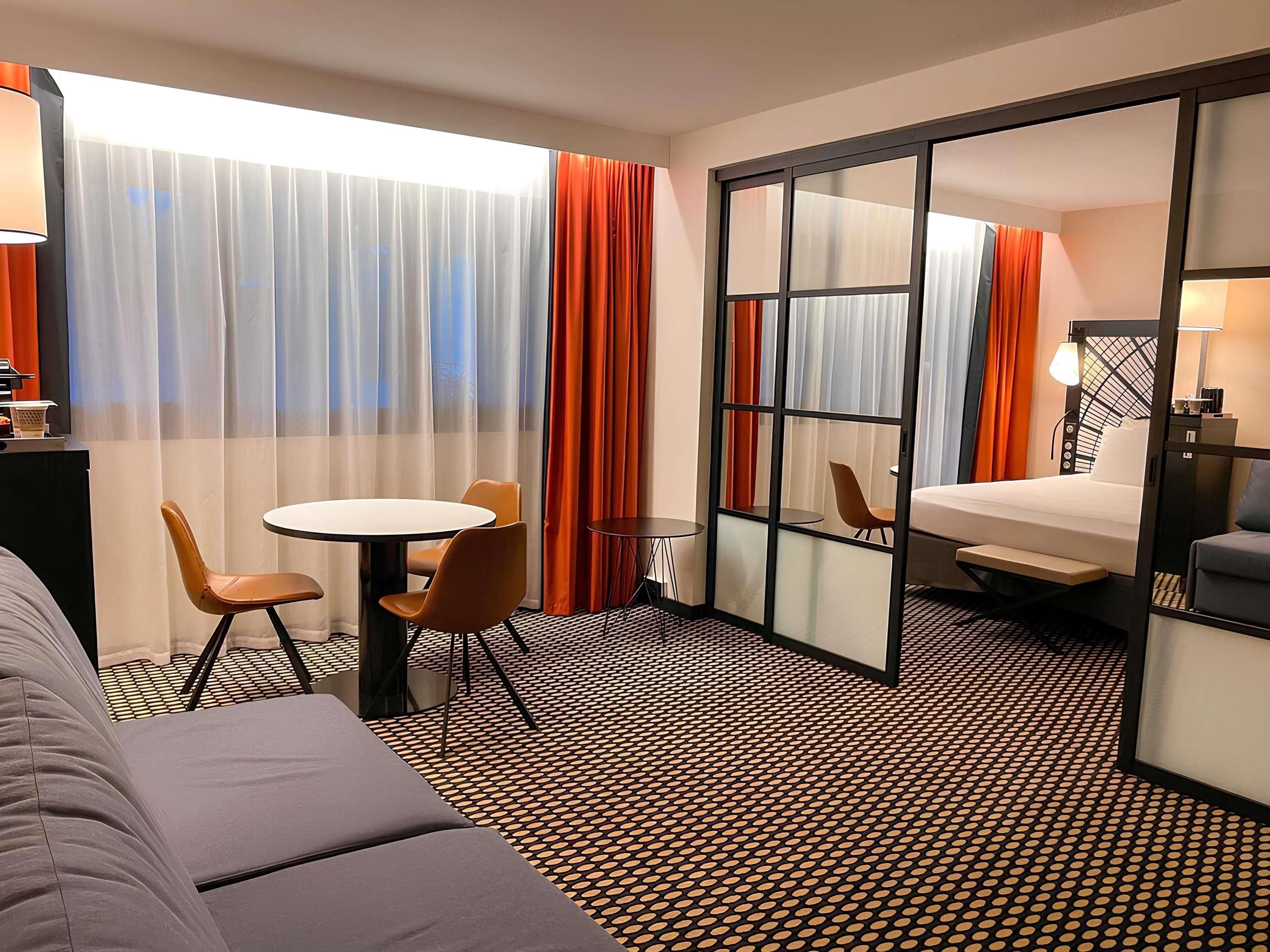 A hotel room with a bed, a sofa, a dining table with chairs, a small table with a chair, and a large window with curtains.
