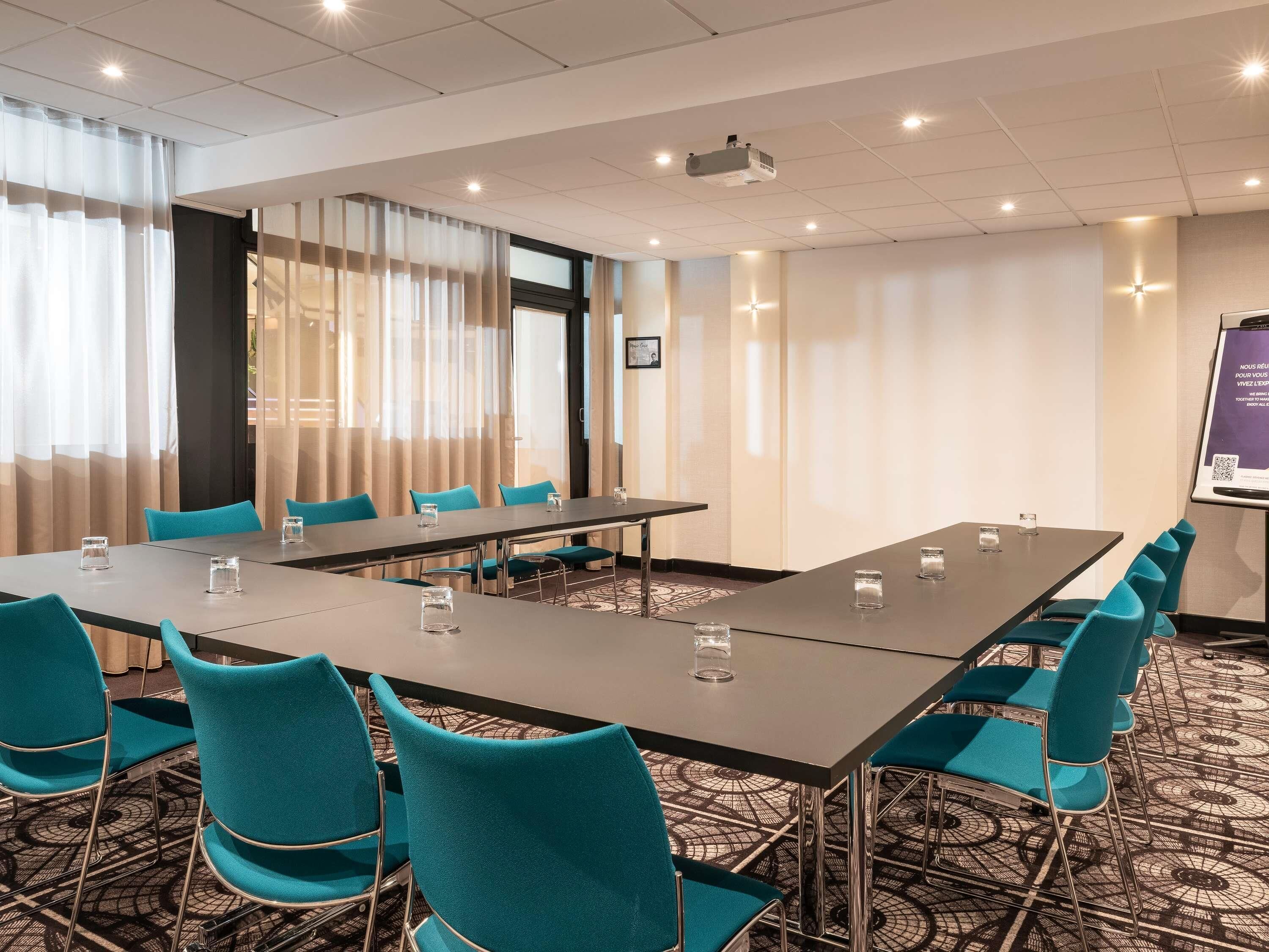 Uma sala de conferências com uma mesa comprida, cadeiras azul-turquesa e uma tela de projetor.