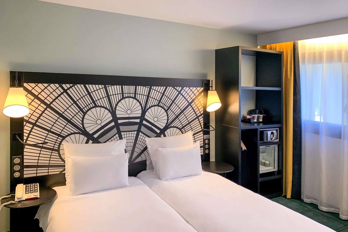 A hotel room with a modern headboard, two beds with white linens, bedside lamps, a minibar, and a view of the outside through sheer curtains.