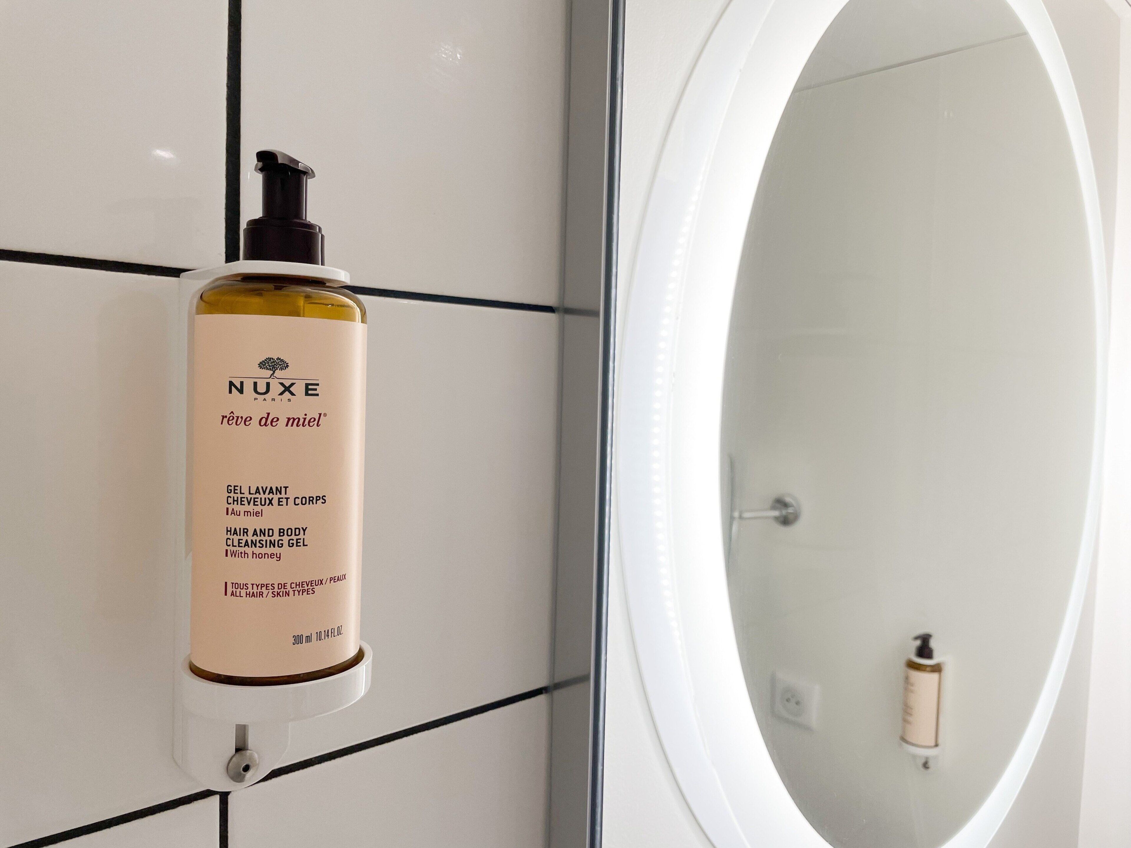A bathroom with a Nuxe shower gel dispenser mounted on a tiled wall and a large oval mirror.