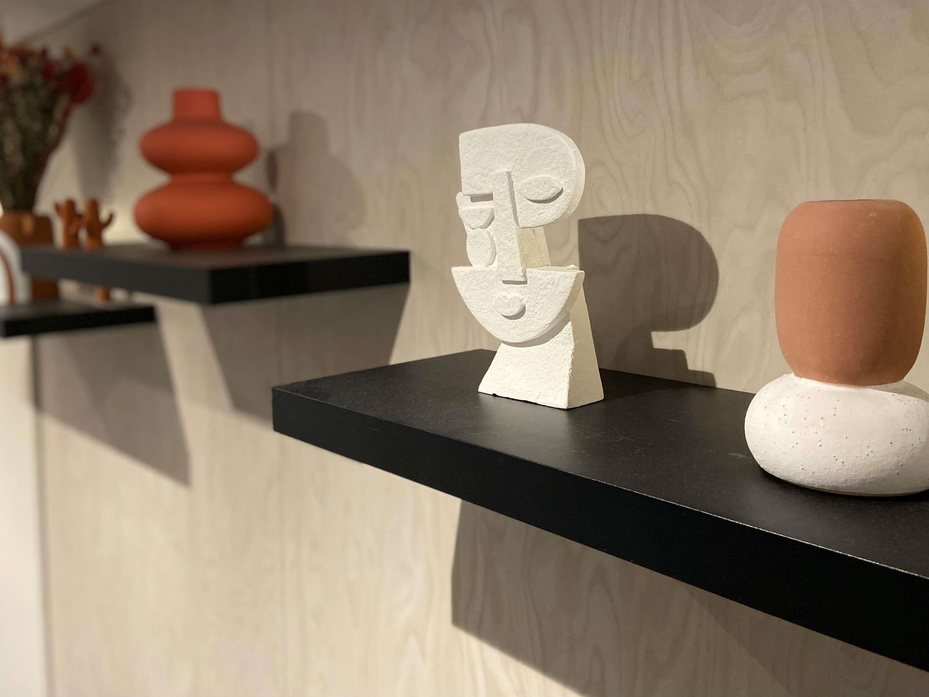 Ein modernes Interieur mit minimalistischer Einrichtung, darunter eine weiße abstrakte Skulptur auf einem schwarzen Regal, eine strukturierte Vase und ein roter Topf an einer Holzwand.