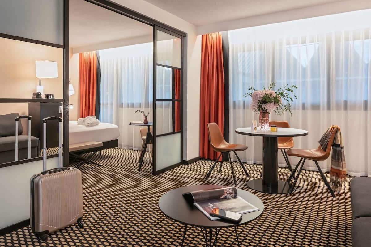 A hotel room with a bed, a desk, a chair, a table, a suitcase, and a magazine.