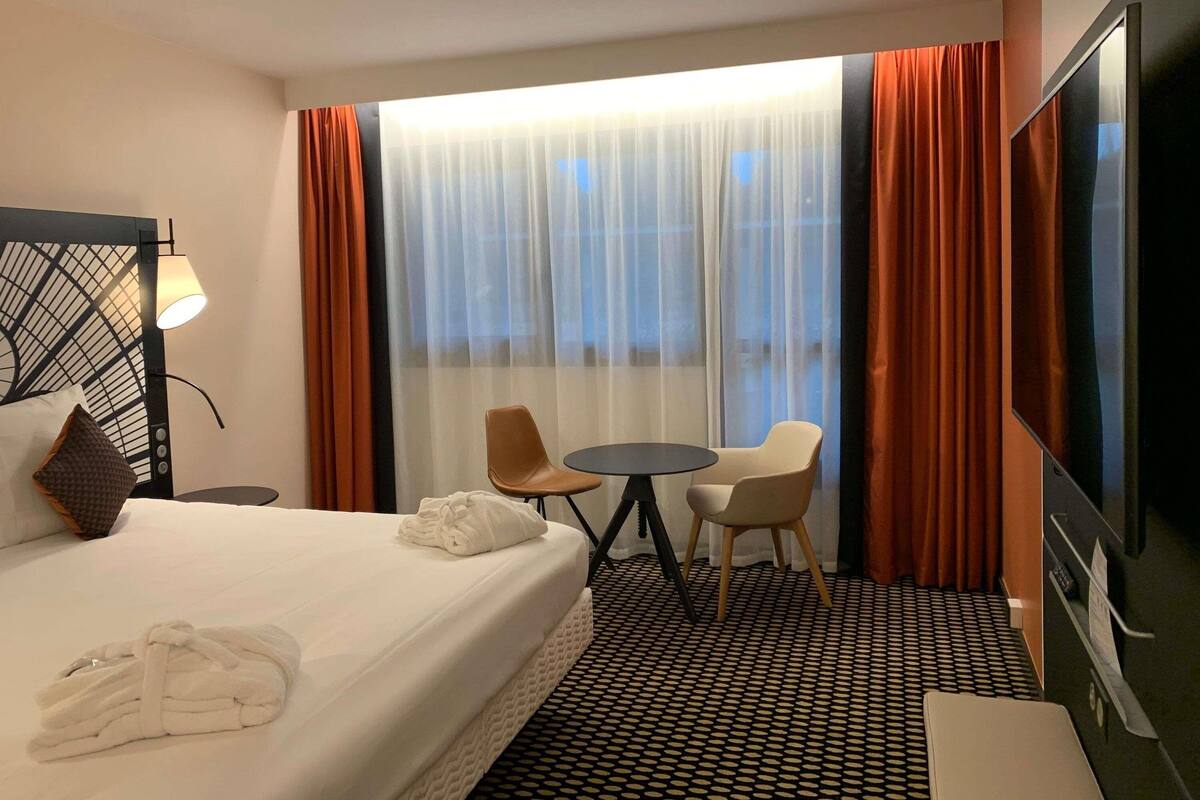 A hotel room with a bed, a desk, a chair, a TV, and a large window with curtains.
