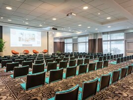 Una sala de conferencias con filas de sillas, una pantalla de proyector y un ventanal grande.