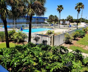 A pool area with a fenced deck, palm trees, and a building with a blue roof in the background. - Days Inn by Wyndham Titusville Kennedy Space Center (Titusville)