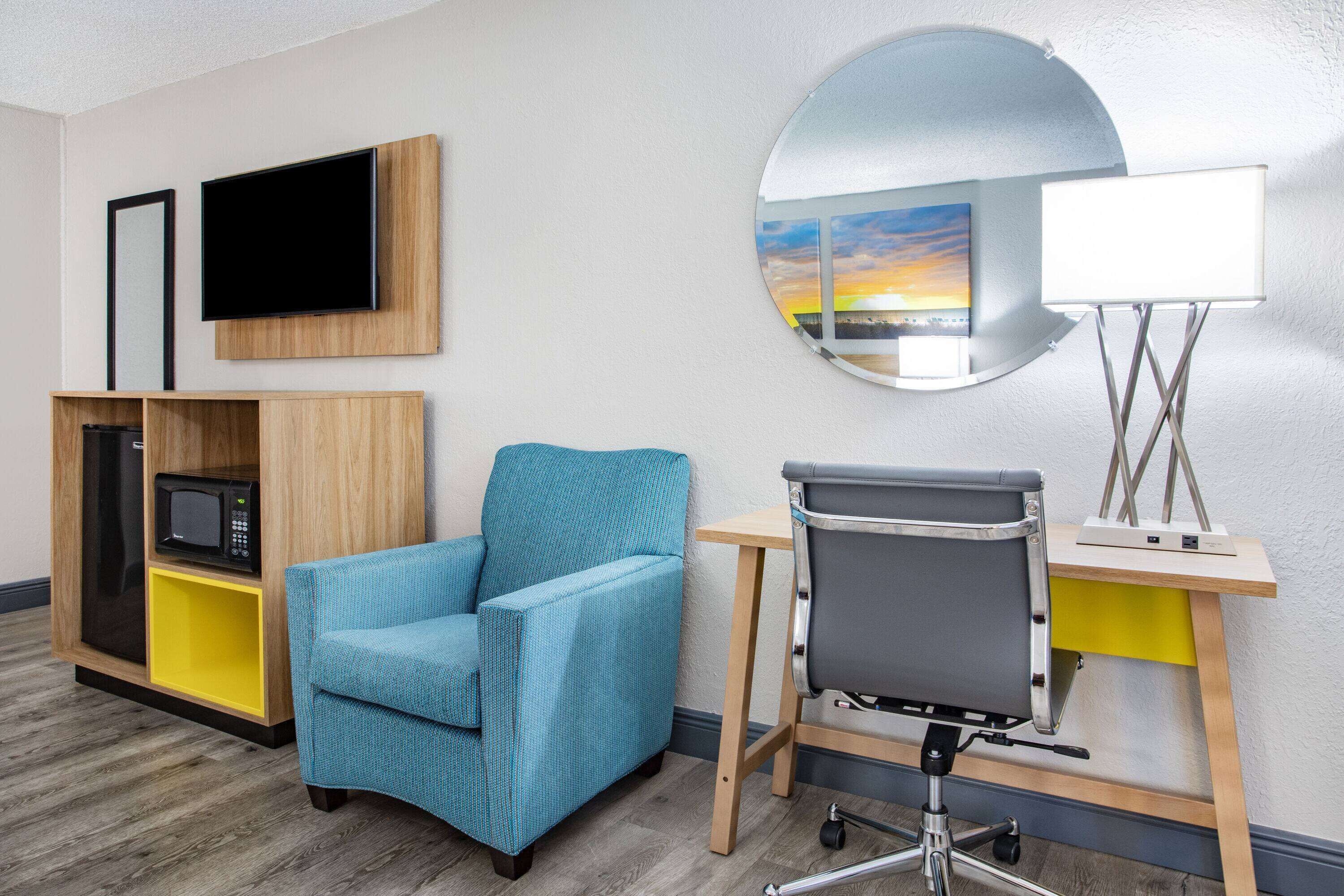 A hotel room with a blue armchair, a desk with a lamp, a TV, and a mirror reflecting a sunset view.