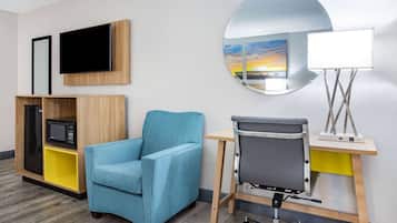 Habitación de hotel con un sillón azul, un escritorio con lámpara, televisor y espejo que refleja una vista al atardecer.