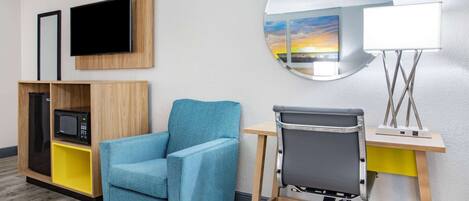 A hotel room with a blue armchair, a desk with a lamp, a TV, and a mirror reflecting a sunset view.