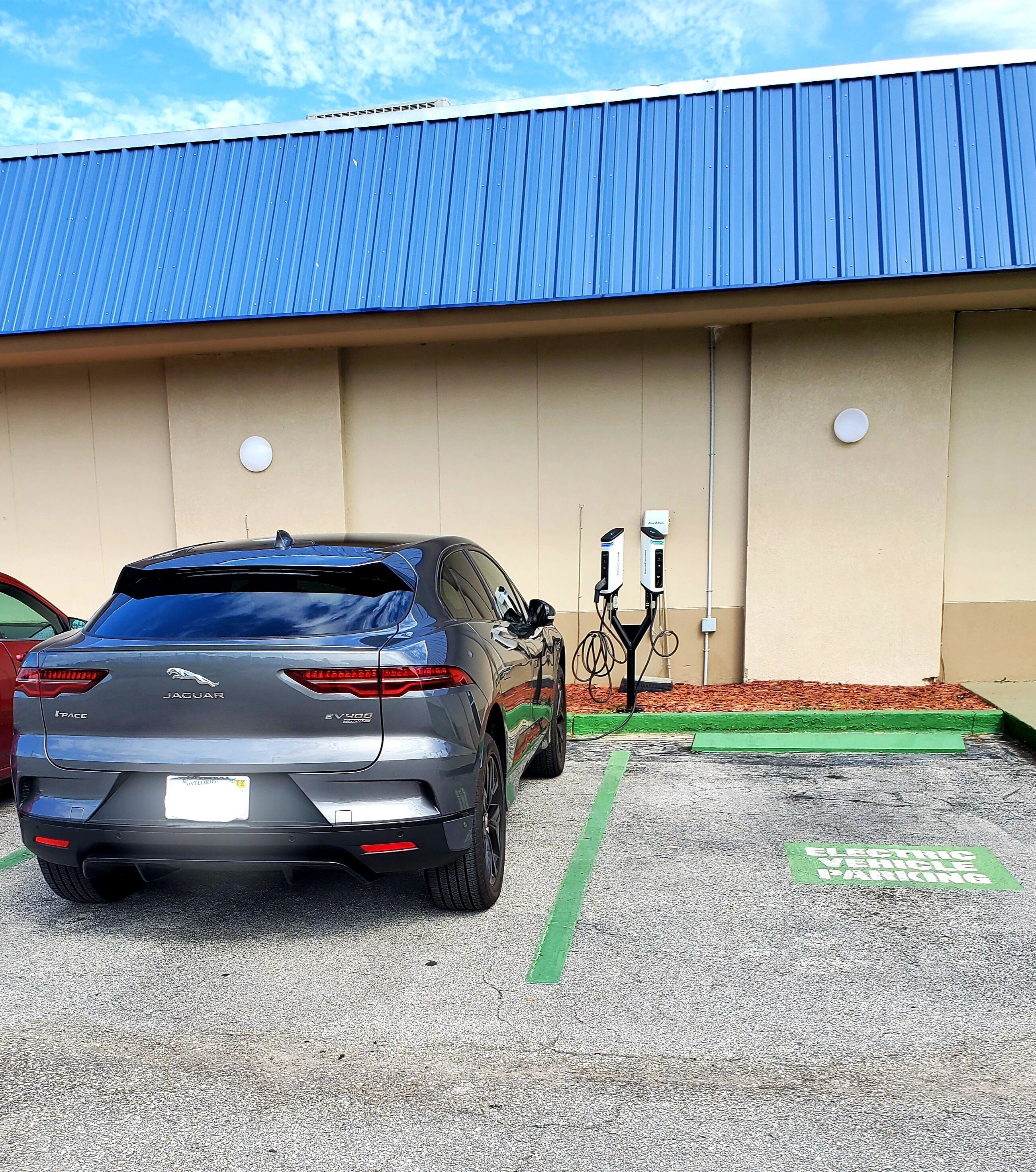 Un área de estacionamiento con un Jaguar I-Pace EV300, una estación de carga y un edificio con techo azul.