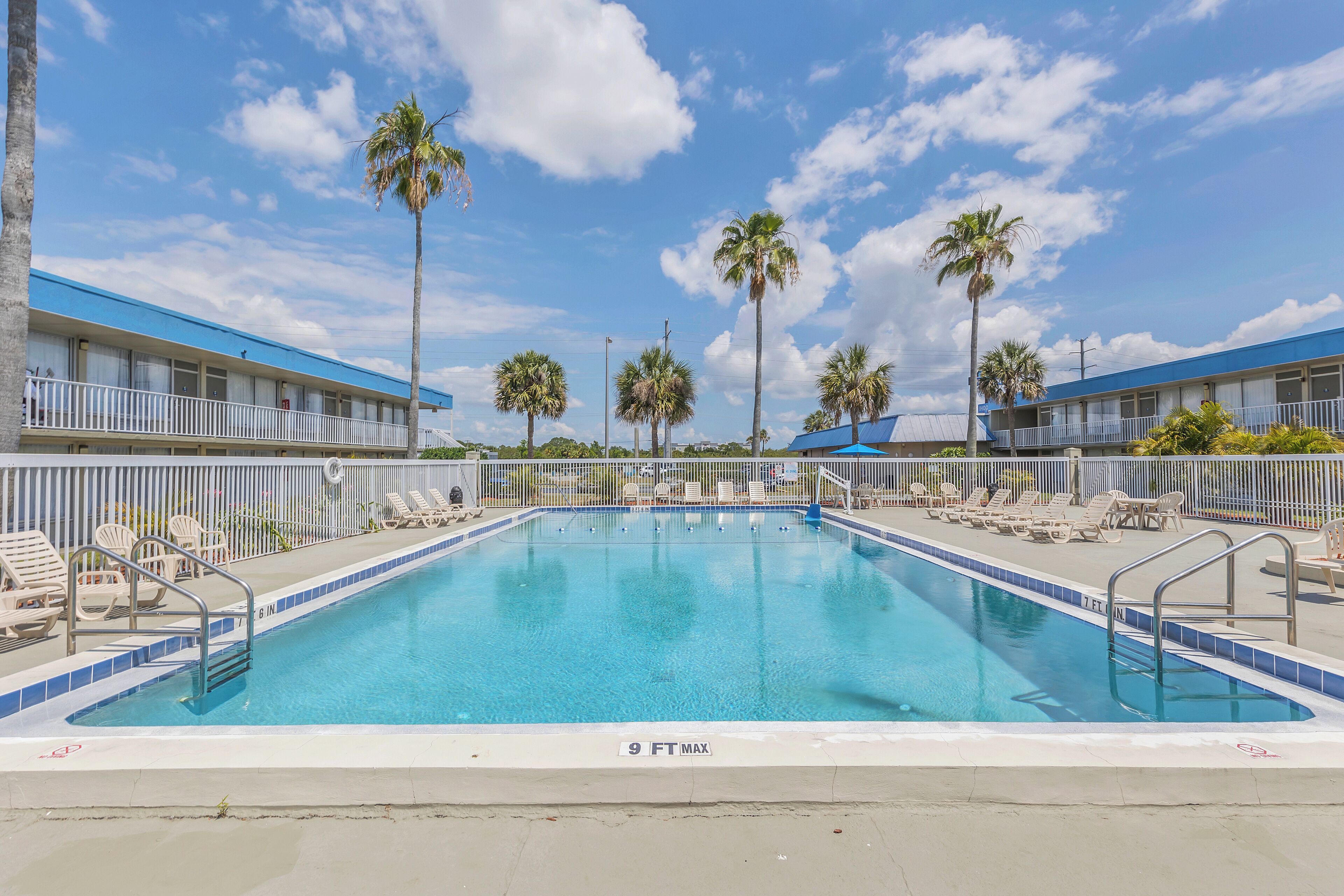 Una piscina con una profundidad máxima de 9 pies, rodeada de sillas de descanso y palmeras, con un edificio de techo azul al fondo.