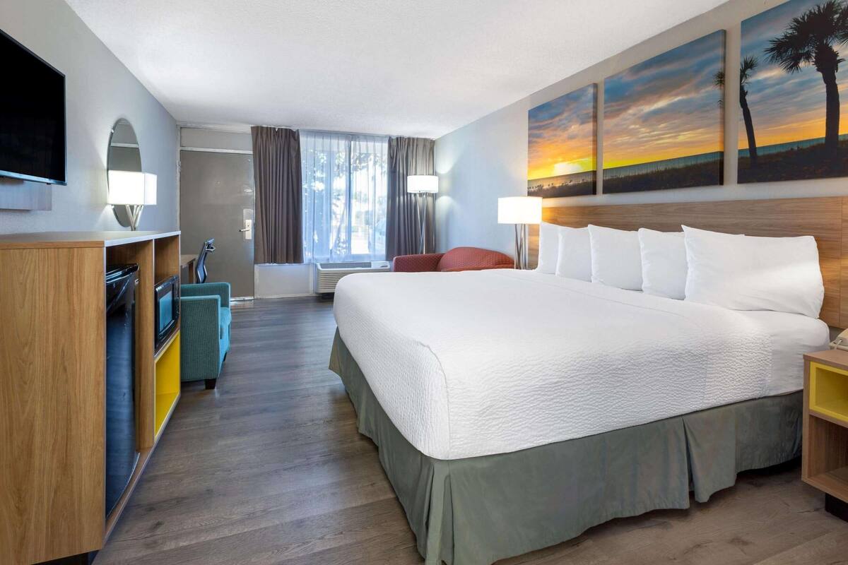 Ein Hotelzimmer mit einem großen Bett, einem Fernseher, einem Schreibtisch, einem Sessel und einem Sonnenuntergangsbild.