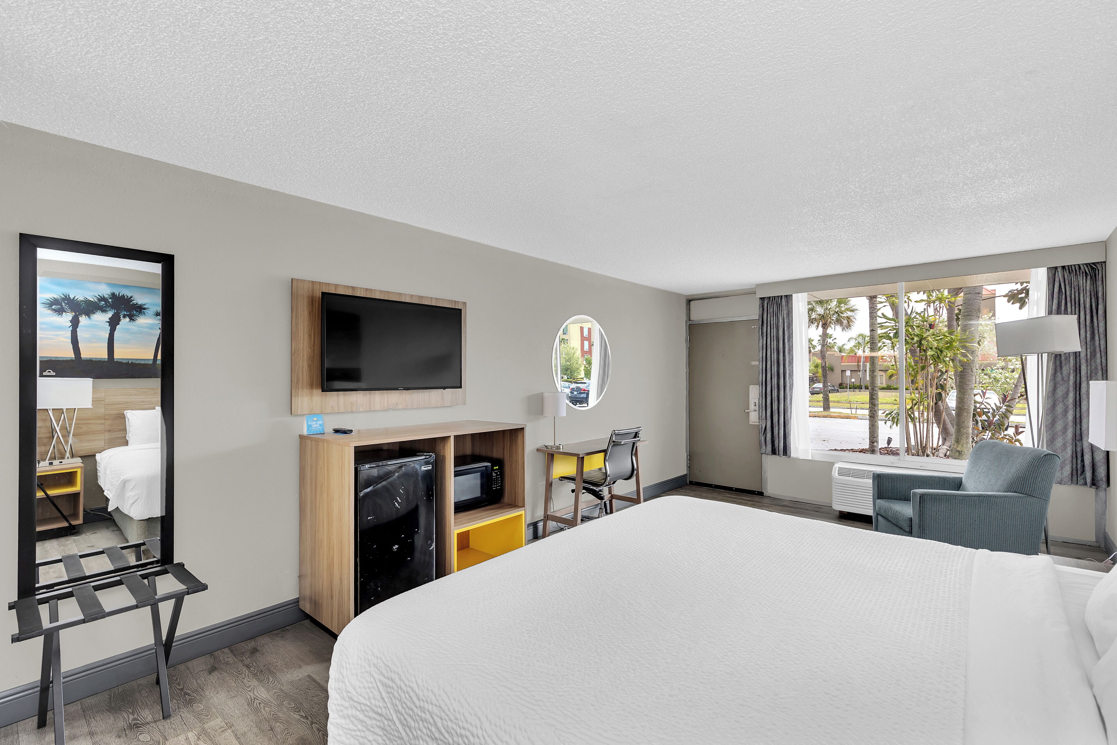 A hotel room with a large bed, a TV, a desk with a chair, and a window with a view of palm trees.