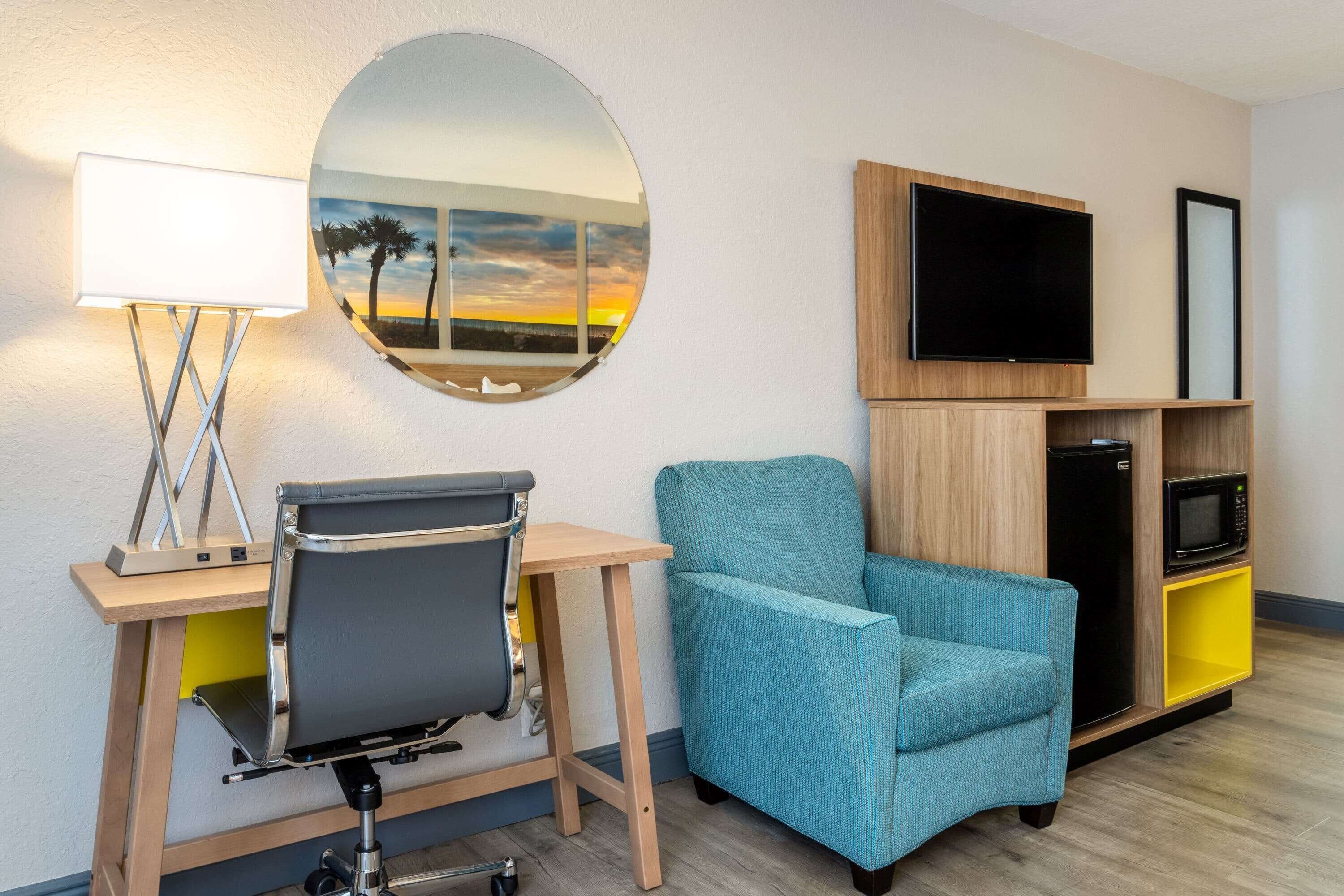 Habitación de hotel con escritorio, silla, televisión, microondas y espejo con vistas al atardecer.