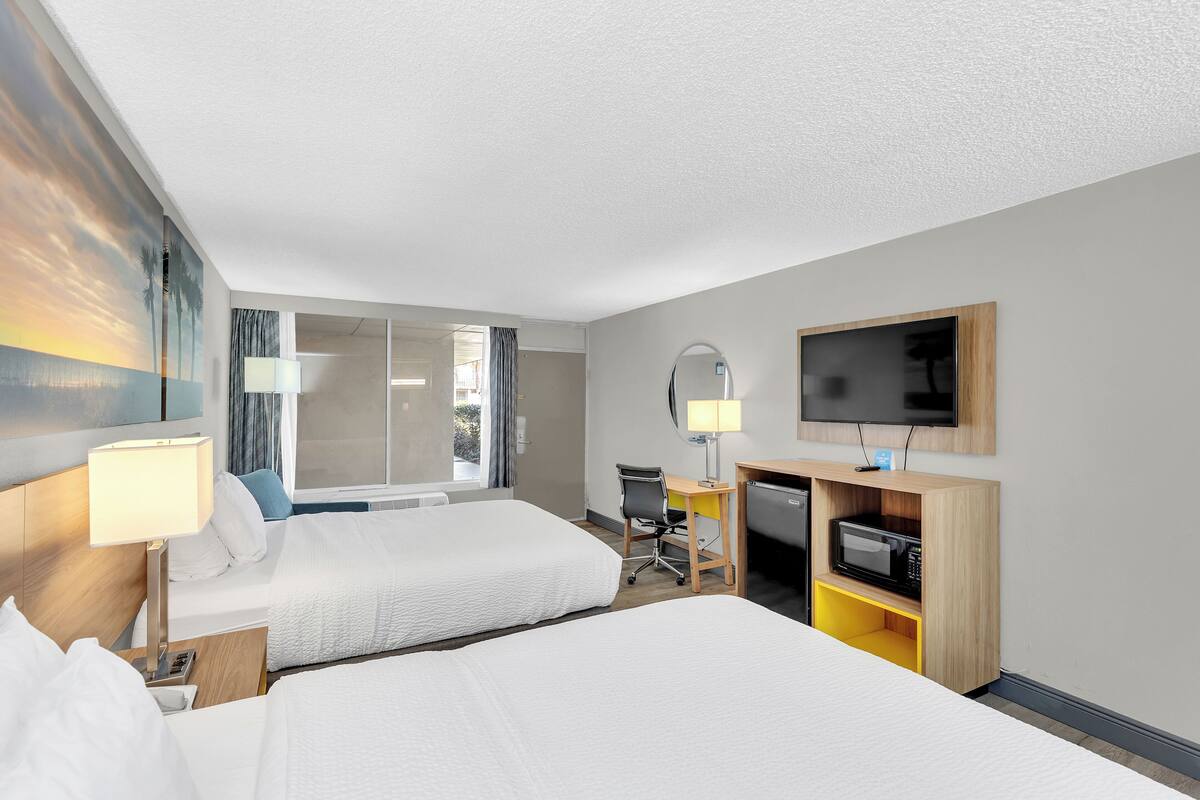 Ein Hotelzimmer mit zwei Betten, einem Schreibtisch mit Stuhl, einem Fernseher und einem Fenster mit Vorhängen.
