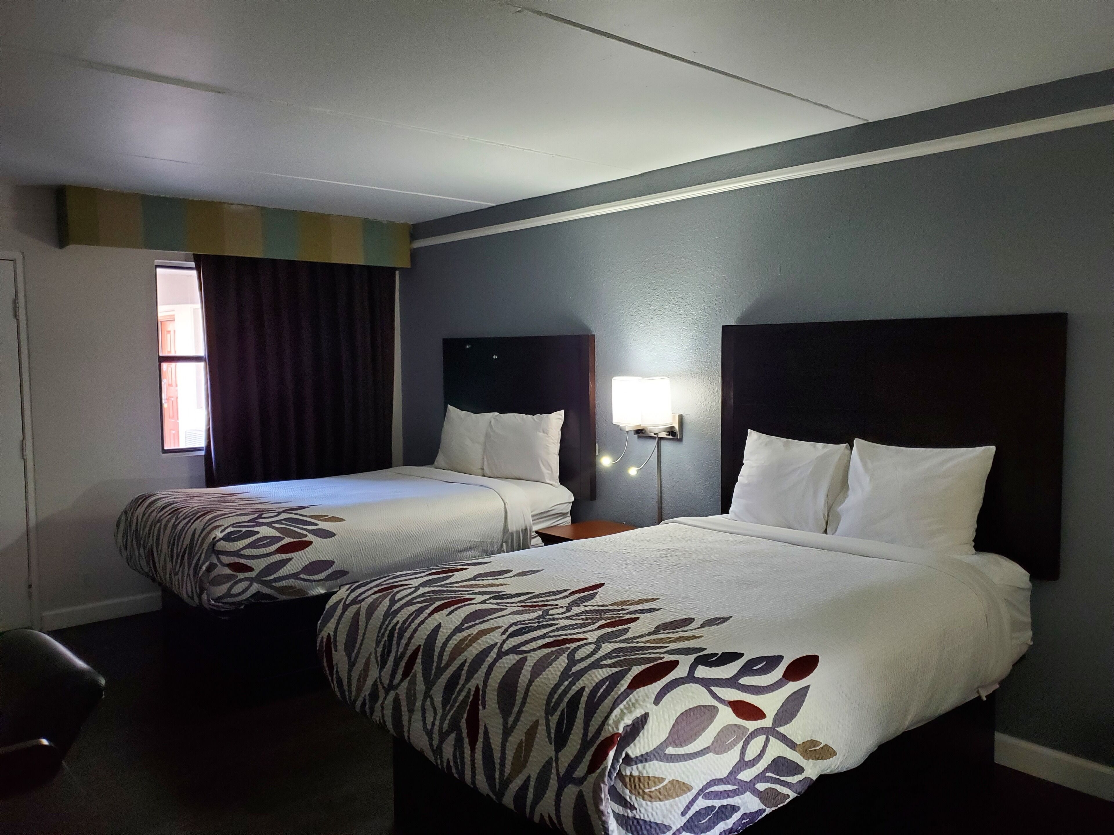 A hotel room with two beds, a window with curtains, a nightstand with a lamp, and a chair.