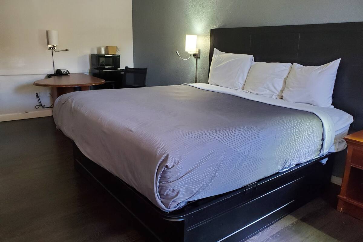 A neatly made bed with a grey comforter and white pillows, a small wooden side table, a wall-mounted lamp, and a television on a small table.