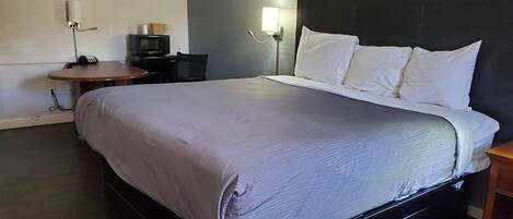 Una cama bien tendida con edredón gris y almohadas blancas, una pequeña mesita de noche, una lámpara de pared y un televisor sobre una mesita.