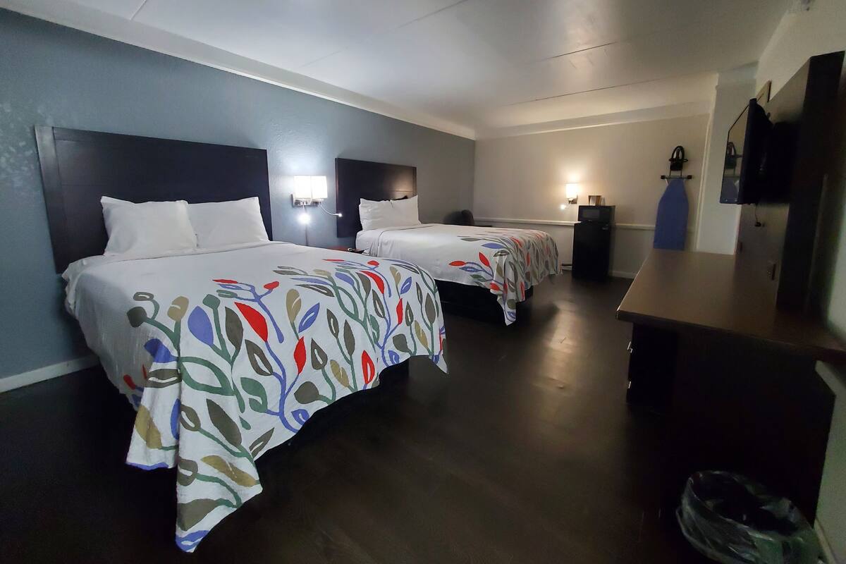 A hotel room with two beds, a desk, a TV, and a wall-mounted light.