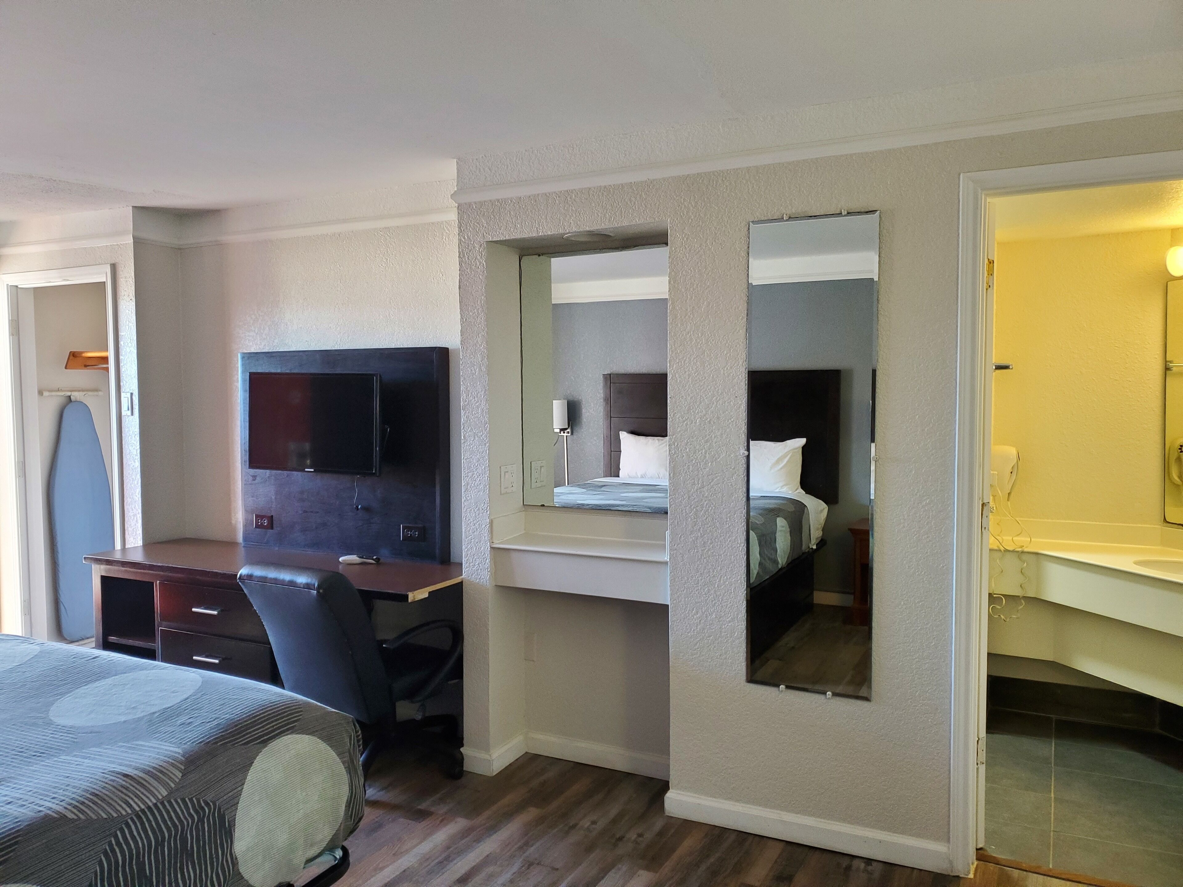 A hotel room with a bed, a desk with a TV, a mirror, and a bathroom with a sink and toilet.
