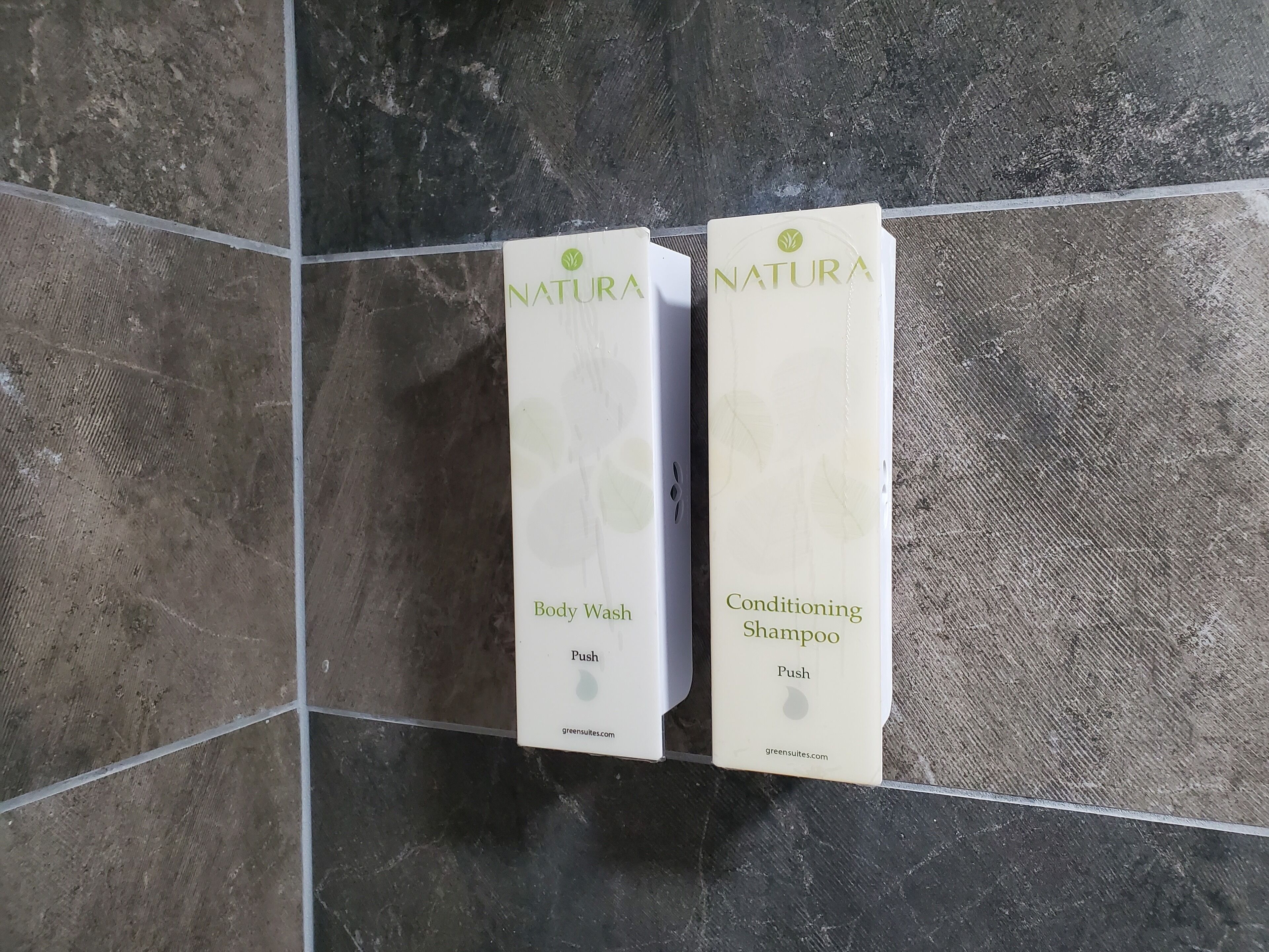Two Natura body wash and conditioning shampoo boxes on a tiled wall.