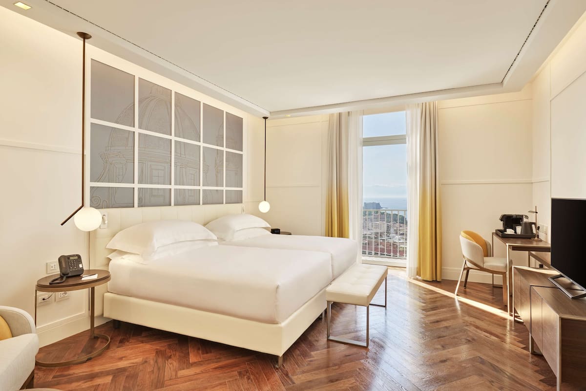 A hotel room with a large bed, a desk with a chair, a TV, and a view of the city through the window.